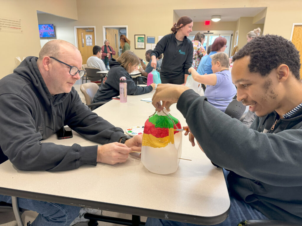 Jim Mahanes of Jewish Family and Career Services sits at a table with a Cedar Lake man painting a milk container into a birdhouse