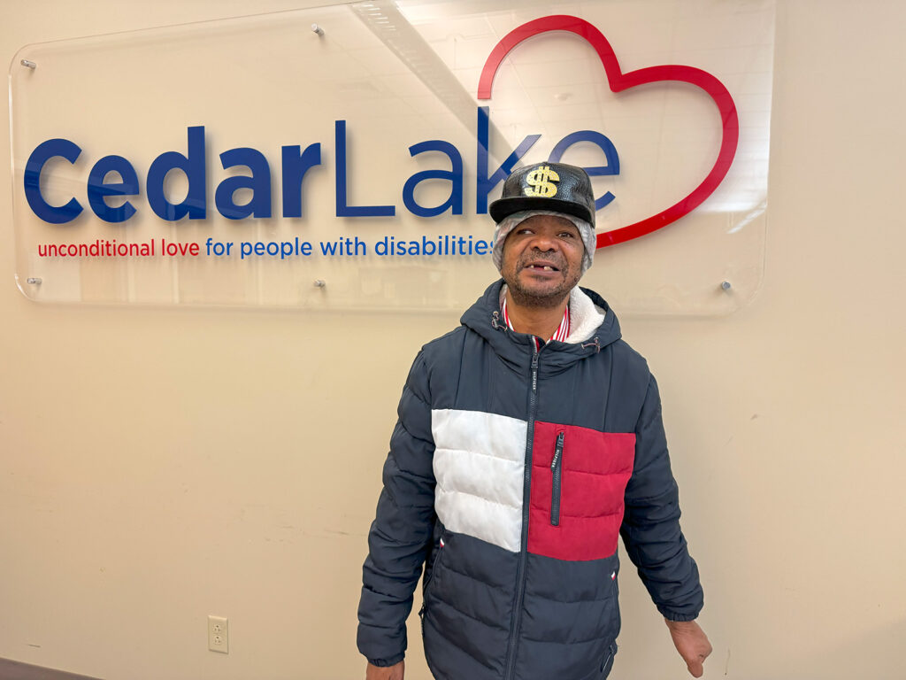 Man smiles wearing a blue hat and puffer jacket in front of a sign that reads Cedar Lake