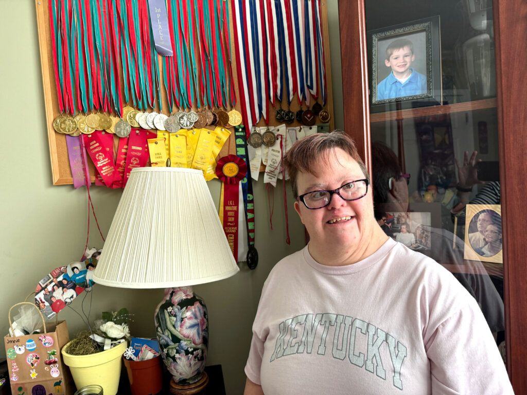 A women in a pink shirt smiles in front of a wall of hanging medals